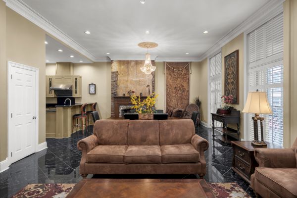 A spacious living room with beige walls, brown sofas, a chandelier, bar area, and decorative elements like art pieces and plants.
