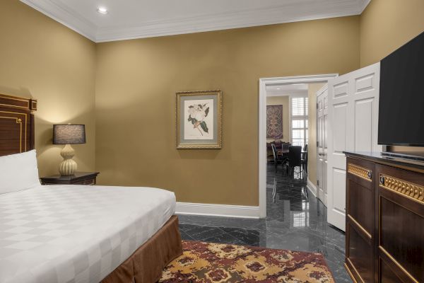 A bedroom with a large bed, nightstand with a lamp, framed art, and a dresser with a TV. An open door leads to another room.