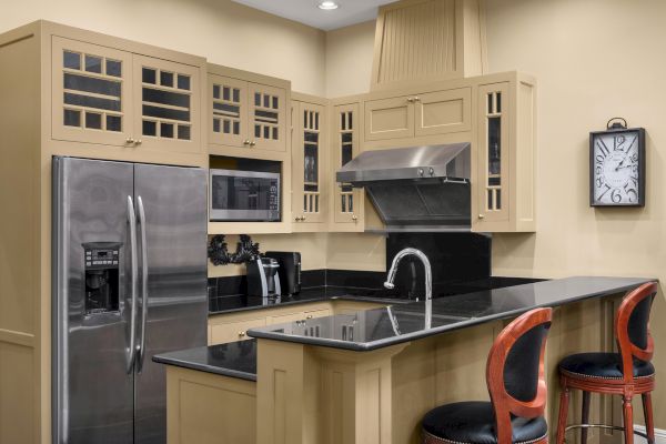 A modern kitchen with beige cabinets, stainless steel appliances, a black countertop, and bar stools. A wall clock hangs on the wall.