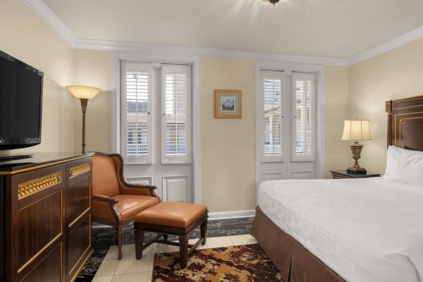 A cozy room with a bed, armchair, TV, and lamps. White shutters cover the windows. Artwork hangs on the beige walls.