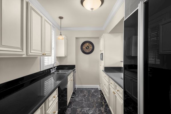 A modern kitchen with cream cabinets, black countertops, a wall clock, pendant light, and black marble floor.