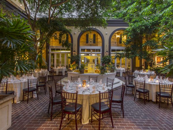 An elegant courtyard set up for dining with round tables, white tablecloths, chairs, soft lighting, and lush greenery around.