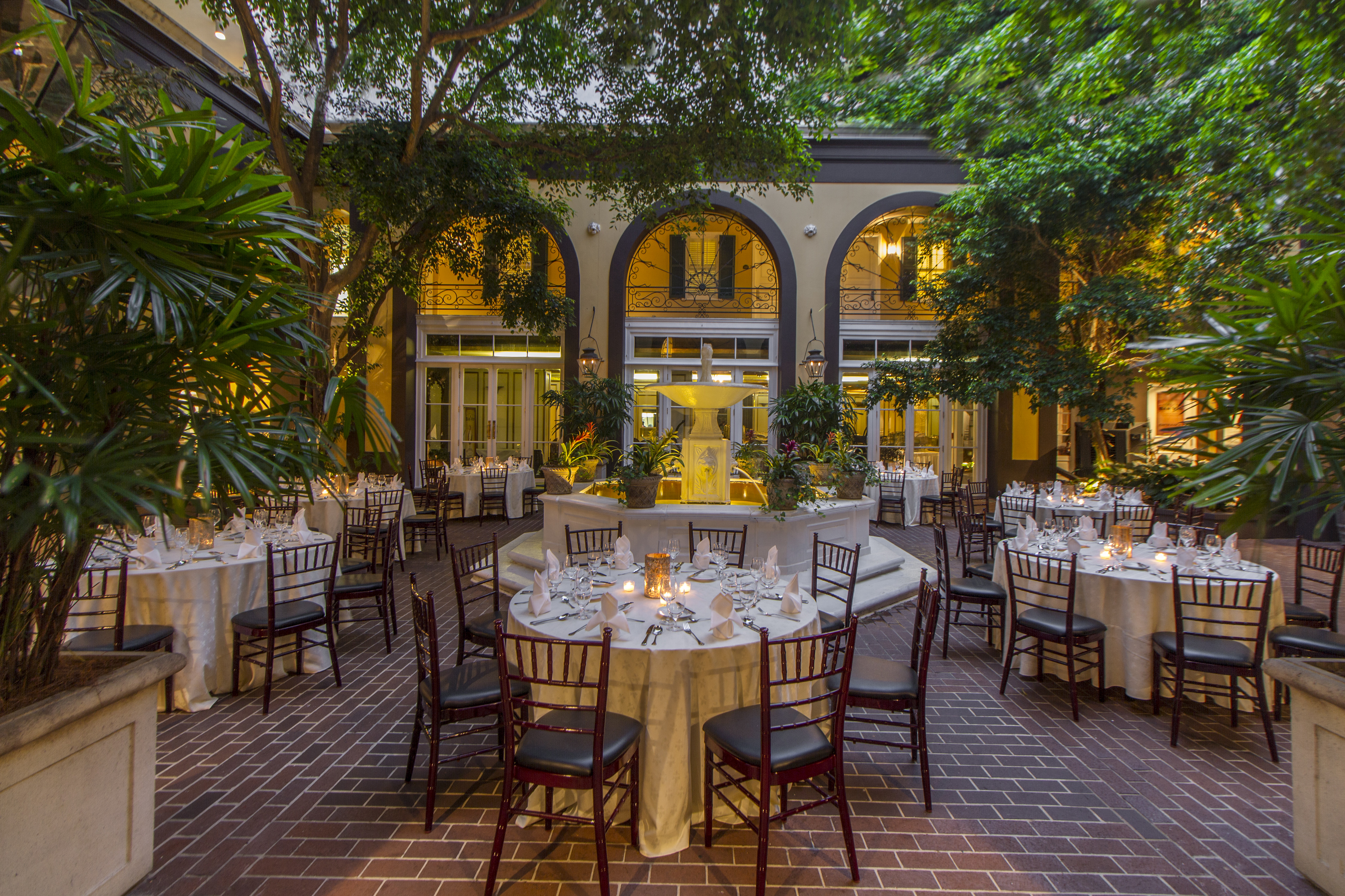 A beautifully set outdoor dining area with round tables, white tablecloths, and dark chairs, surrounded by plants and an archway.