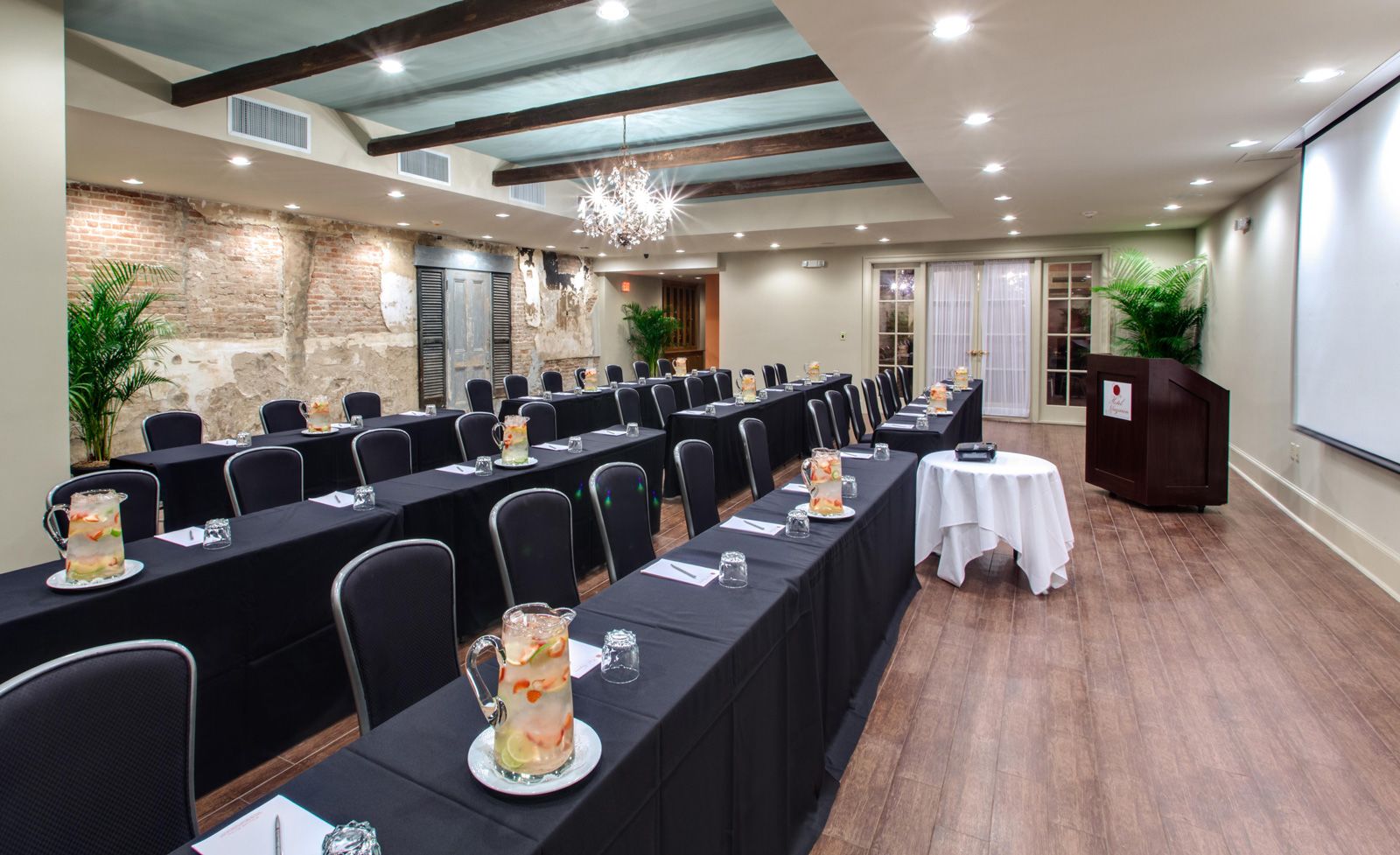 a long, well-lit meeting room with rows of black tables and chairs, pitchers of water, and a projector screen