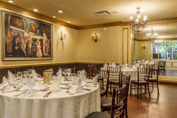 The image shows a dining room with round tables set for an event, adorned with a painting and elegant lighting.