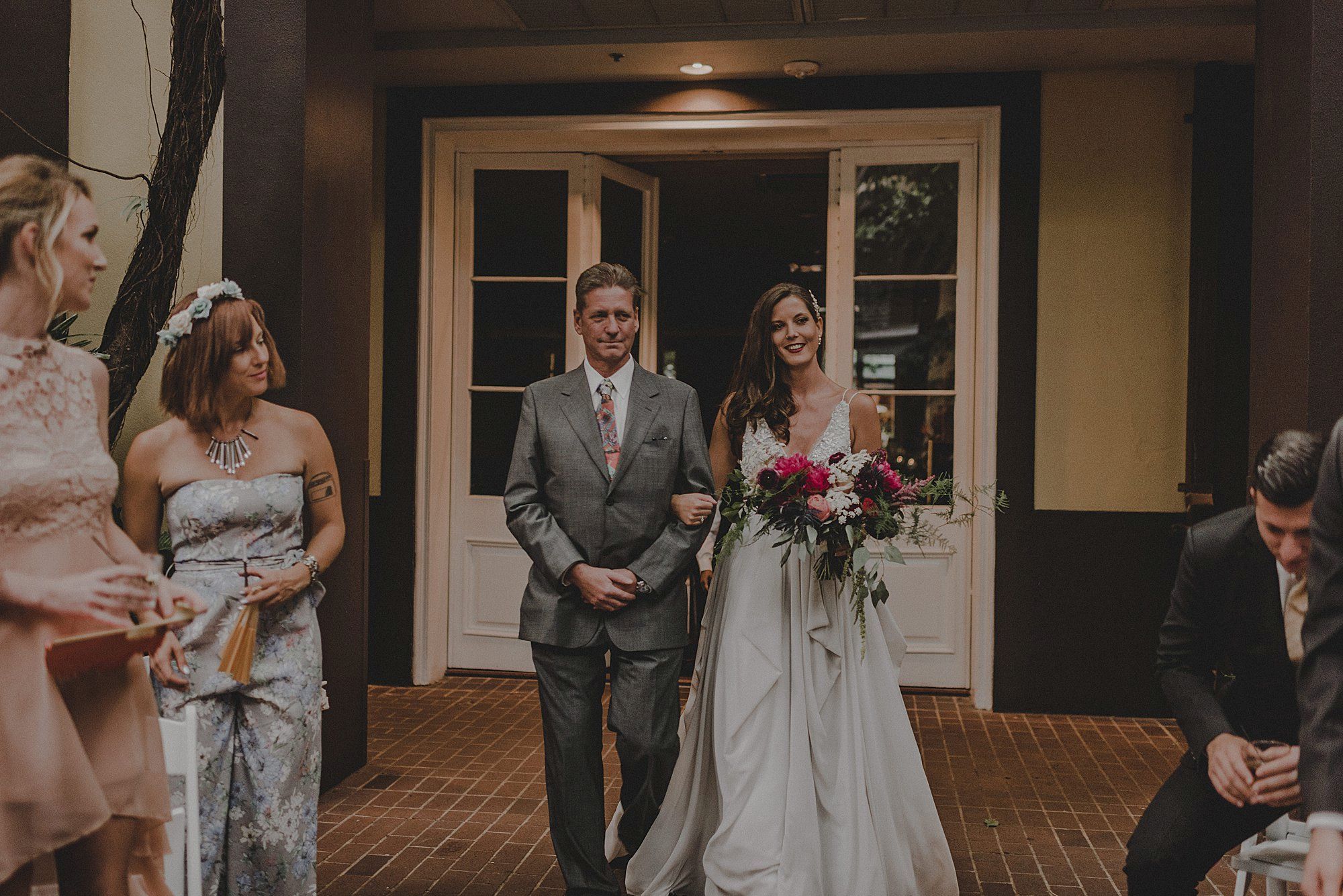 A bride in a white dress walks down the aisle with a man, holding flowers, surrounded by elegantly dressed guests at a wedding venue.