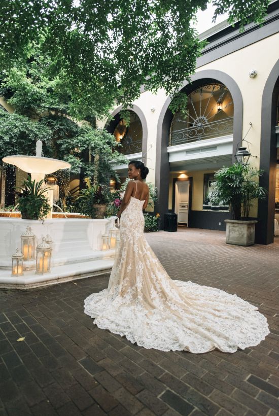 A woman in a bridal gown stands by a fountain in an elegant courtyard, surrounded by greenery and lanterns.