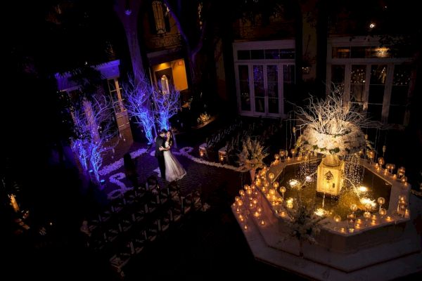 A couple dances in a beautifully lit outdoor venue with candles and a decorated fountain, creating a romantic atmosphere.