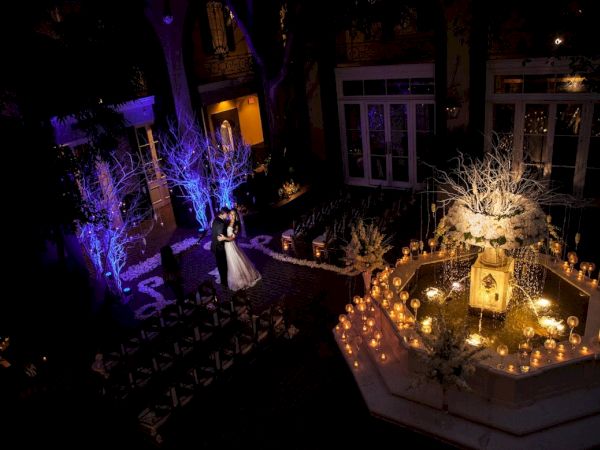 A couple dances in a beautifully lit outdoor venue with candles and a decorated fountain, creating a romantic atmosphere.