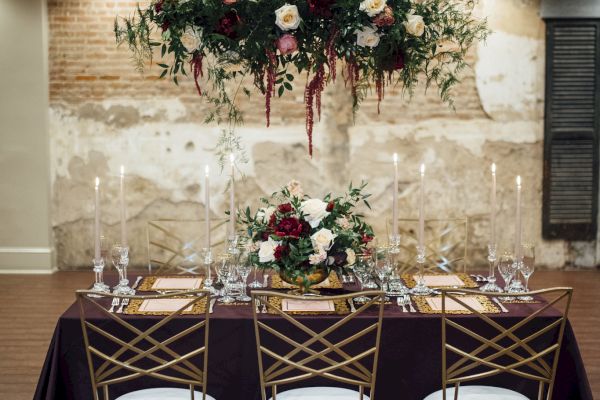 Elegantly set dining table with floral arrangements, tall candles, a chandelier, and gold chairs against a rustic brick wall backdrop.