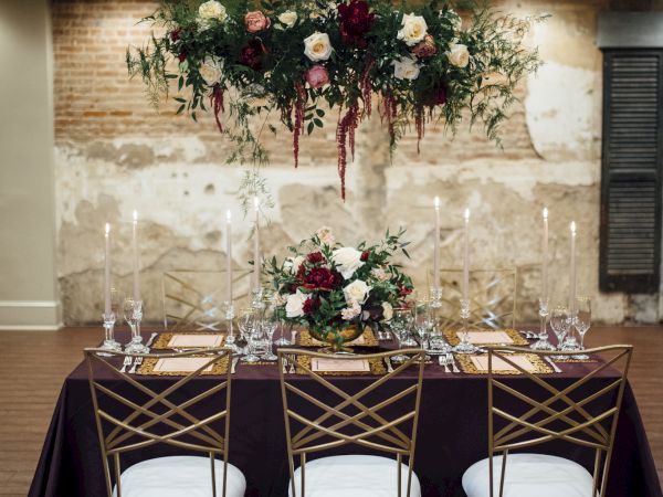 Elegantly set dining table with floral arrangements, tall candles, a chandelier, and gold chairs against a rustic brick wall backdrop.