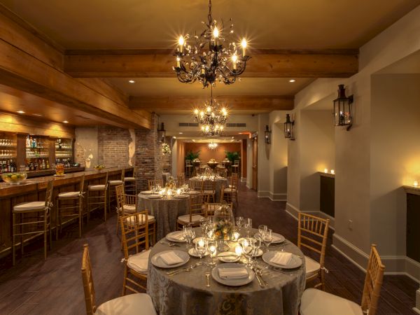 A warmly lit, elegant dining area with chandeliers, set tables, and a wooden bar, creating a cozy atmosphere.