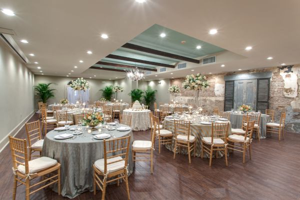 The image shows a decorated banquet hall with elegantly set tables, floral arrangements, and gold chairs, ready for an event or celebration.