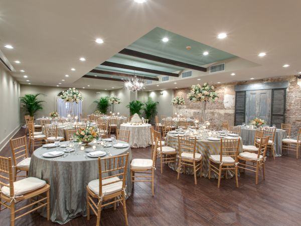 The image shows a decorated banquet hall with elegantly set tables, floral arrangements, and gold chairs, ready for an event or celebration.