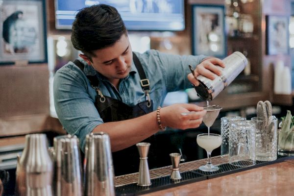 A bartender in a denim shirt and apron pours a cocktail through a strainer into a glass at a bar counter.