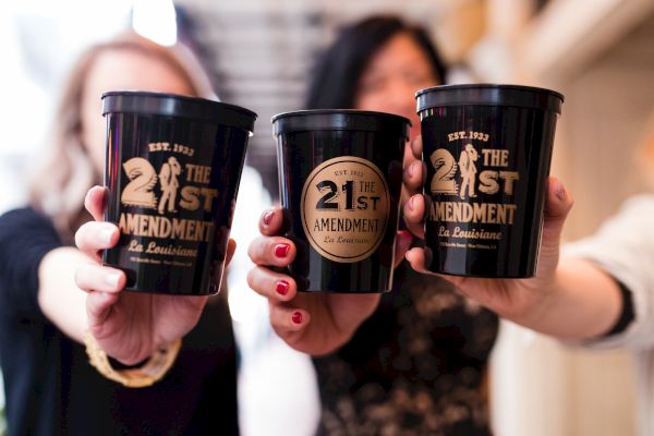 Three people holding black cups labeled "21st Amendment" from a place called La Louisiane, established in 1933.