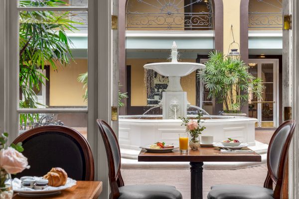 A dining area with breakfast on the table, elegant chairs, a fountain outside, and lush greenery in an open courtyard setting.