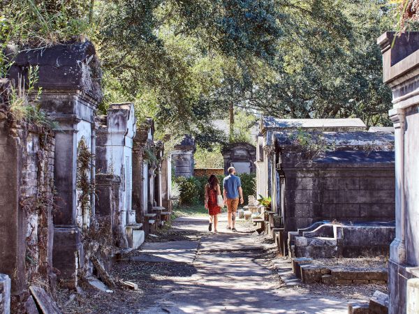 A couple is walking through a historic cemetery with old stone tombs and trees lining the path.
