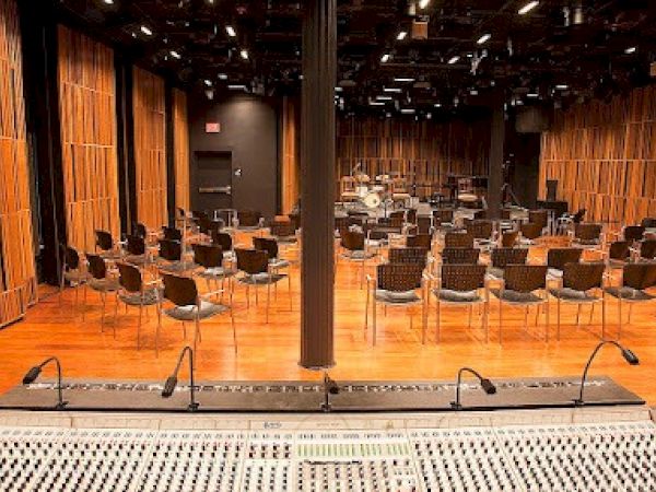 The image shows a recording studio with a mixing console in the foreground and rows of chairs with a drum set in the background.
