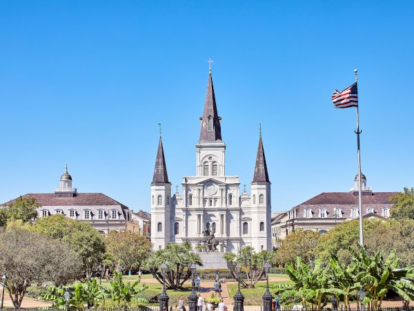 The image shows St. Louis Cathedral in New Orleans, with a clear blue sky and a U.S. flag flying nearby within a garden area.