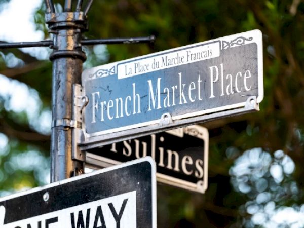 A street sign reads "French Market Place" with another sign partially visible and a "One Way" sign below, surrounded by trees.