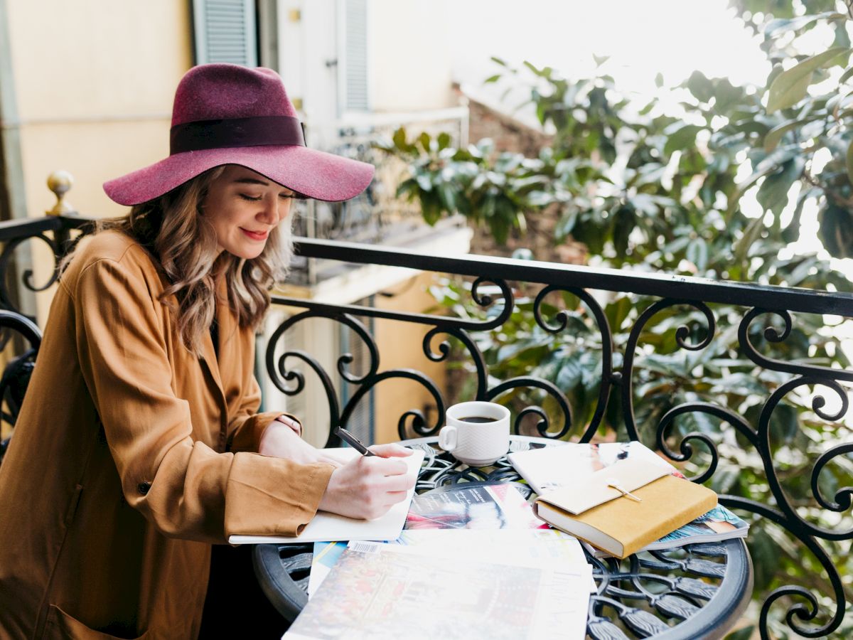 A woman in a wide-brimmed hat writes in a notebook on a balcony with a coffee, surrounded by greenery and books on the table.