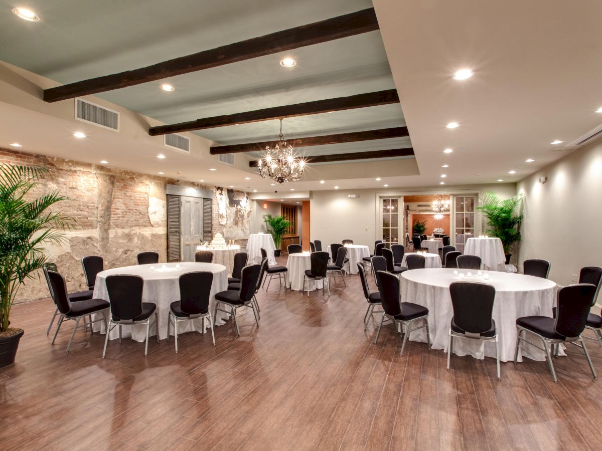 A banquet hall with round tables, white tablecloths, black chairs, chandeliers, potted plants, and a rustic brick wall accent.
