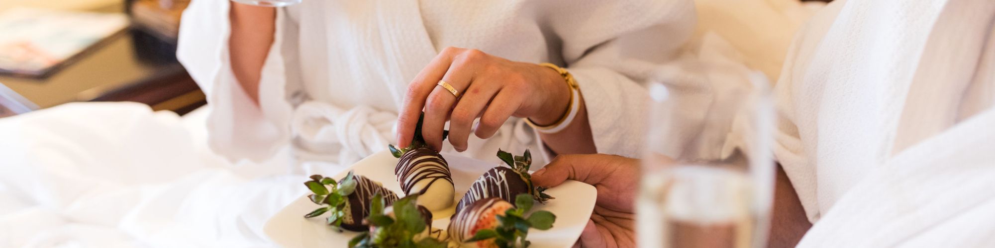 Two people in robes are enjoying chocolate-covered strawberries and champagne in bed.