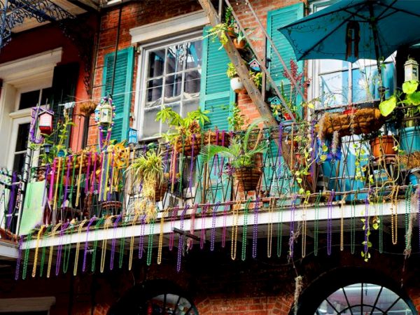 A vibrant balcony with hanging Mardi Gras beads, potted plants, and colorful turquoise shutters, capturing festive charm.