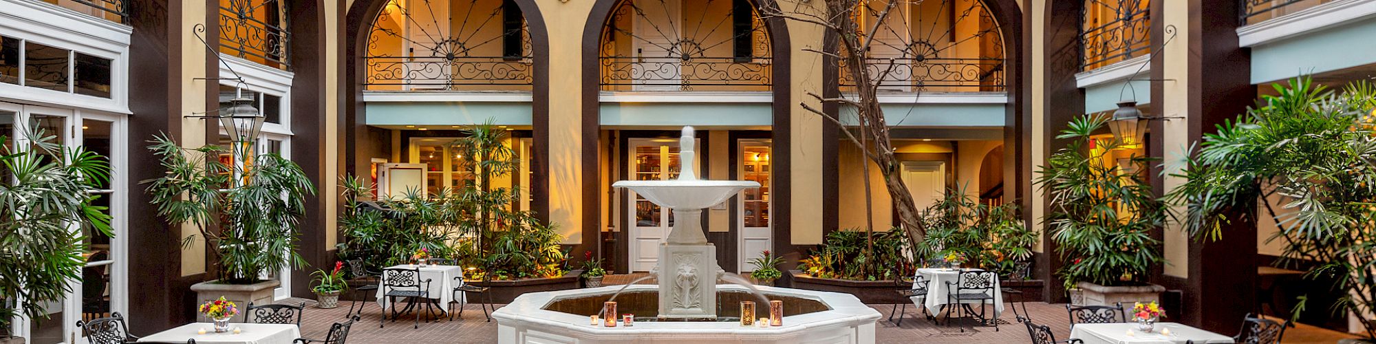A charming courtyard with a central fountain, surrounded by archways, potted plants, and tables with chairs, under a white canopy.