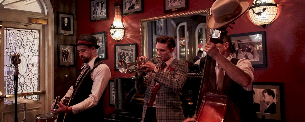 Three men playing musical instruments in a dimly lit room with red walls and framed photos.