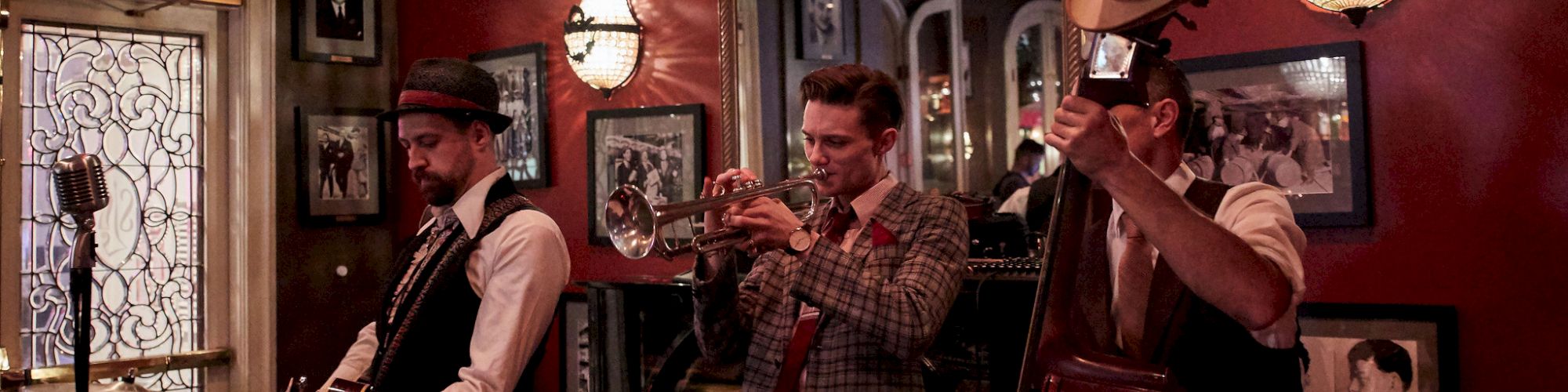 A jazz band with three musicians playing guitar, trumpet, and double bass performs in a dimly lit vintage-themed room.