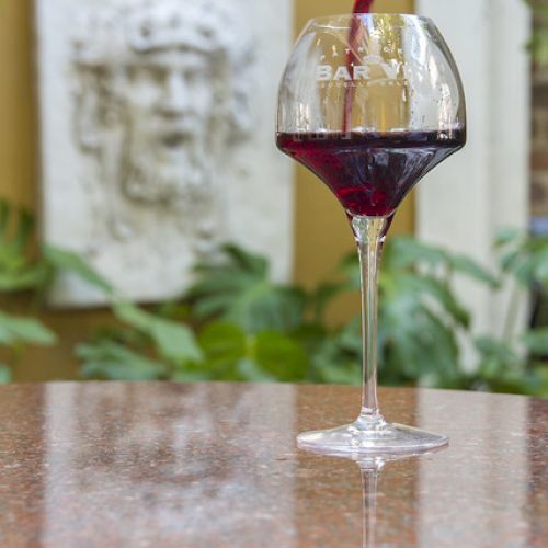 A glass of red wine is being poured on a table, with a stone wall sculpture in the background.
