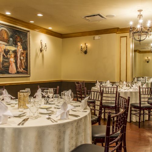 An elegant dining room with round tables set for a formal event, featuring white tablecloths, napkins, wine glasses, and a large painting on the wall.
