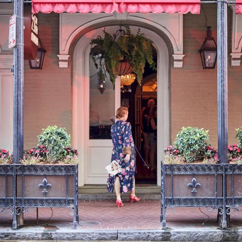 A person in a floral dress enters a building with red awnings, surrounded by planters and a brick walkway.