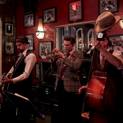 A jazz band performs in a dimly lit room, featuring a guitarist, trumpeter, and bassist. The walls are adorned with framed photos.