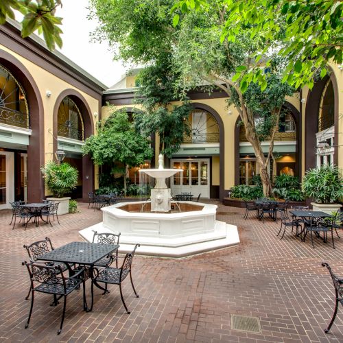 A charming courtyard with a central fountain, surrounded by wrought iron tables and lush greenery beneath a canopy of trees.