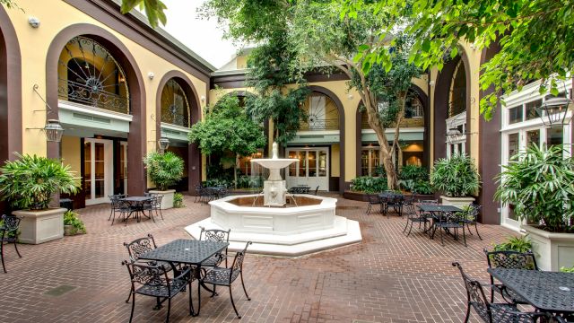 A courtyard with a fountain, outdoor seating, and plants surrounded by a building with arches.