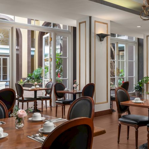 A stylish café interior with elegant wooden tables, chairs, and table settings, featuring large windows and intricate wall decor.