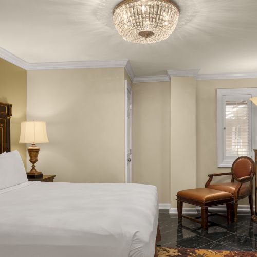 A hotel room with a bed, nightstand, lamp, TV on a cabinet, and chair, all under a chandelier on a tiled floor.