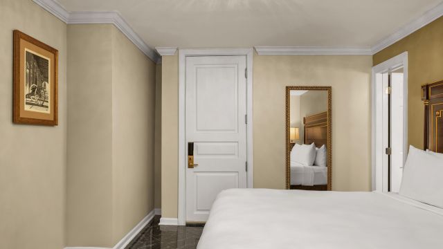 A wide view of a hotel room with a white bed, tan walls, a white door, and a gold-framed mirror.
