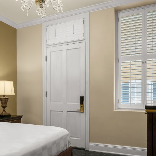 A bedroom with a bed, lamp, wooden furniture, TV, window with shutters, and chandelier, all with beige walls and white trim.