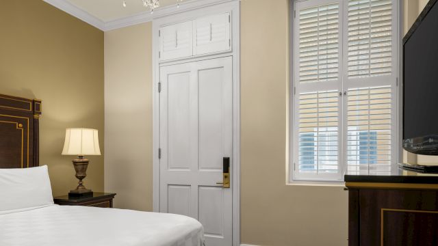 A hotel room with a bed, lamp, window, TV, and door.