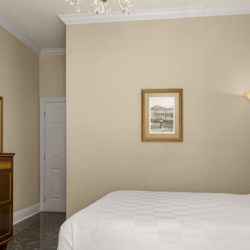 A bedroom with a double bed, TV on a cabinet, tall lamp, framed picture on the wall, and a closed door in a minimalistic setting.