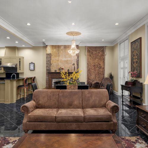 A luxurious living room with a brown sofa, matching armchair, wooden furniture, chandelier, and a view of the kitchen in the background.