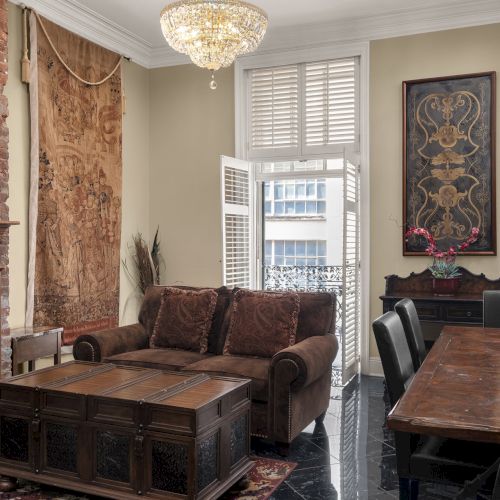 A cozy living room features a brown sofa, wooden table, elegant chandelier, and art on the walls, with large windows allowing natural light.