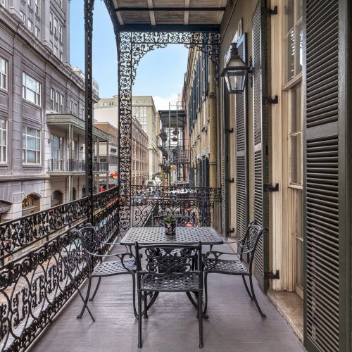 A wrought-iron balcony overlooks a street with historic buildings, featuring a table and chairs, evoking a charming, classic atmosphere.