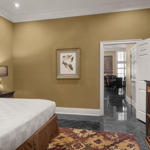 A hotel room with a bed, nightstand, lamp, artwork, TV on a dresser, and a view into another room through an open door.