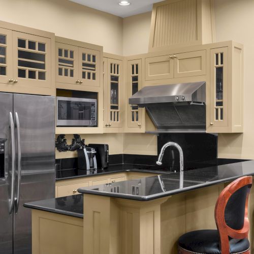 A modern kitchen with beige cabinets, stainless steel appliances, a clock on the wall, and two chairs at the counter.