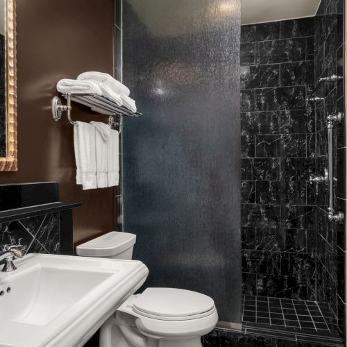 A modern bathroom with a sink, mirror, toilet, and black tiled shower. Towels are neatly placed on the wall rack above the toilet.